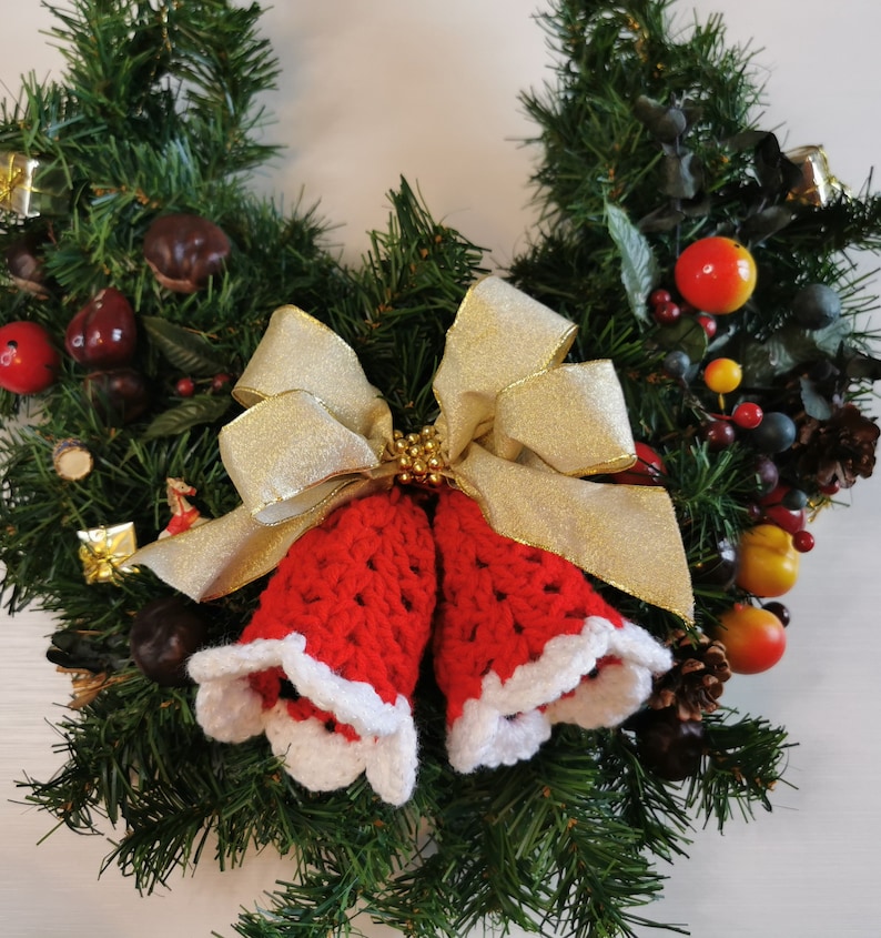 May include: A Christmas wreath decorated with red and white crocheted bells, a gold bow, and various ornaments, including red apples, berries, and pine cones.