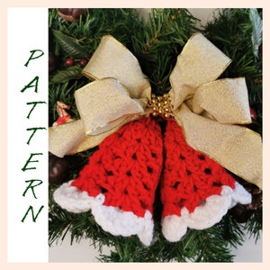 May include: Two red crocheted bells with white trim, tied together with a gold ribbon bow. The bells are hanging on a green Christmas wreath.