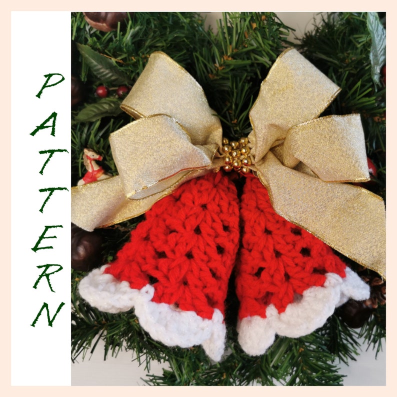 May include: Two red crocheted bells with white trim, tied together with a gold ribbon bow. The bells are hanging on a green Christmas wreath.