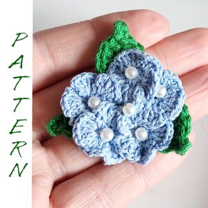 May include: A crocheted flower brooch with blue petals and green leaves. The flower has white pearl beads in the center of each petal.