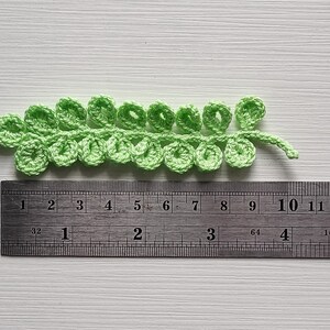 May include: A green crocheted leaf measuring approximately 10 cm long. The leaf is made up of a series of small, connected loops.