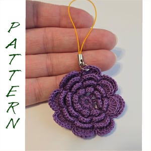 May include: A crocheted purple flower charm with a yellow string and silver clasp. The flower has multiple layers of petals and is held in a hand. The word "PATTERN" is written vertically in green letters.