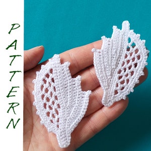 May include: Two white crocheted leaf shapes, with intricate designs, are held in a hand against a turquoise background.
