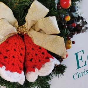 May include: Two red crocheted Christmas bells with white trim, tied with a gold ribbon and gold beads, on a green wreath with red and yellow berries.