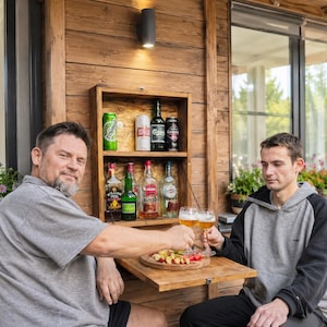 May include: A wall-mounted wooden bar cabinet with two shelves holding various glass bottles and aluminum cans. The cabinet features a fold-down door that serves as a small table surface, currently holding a wooden platter with sliced fruit.