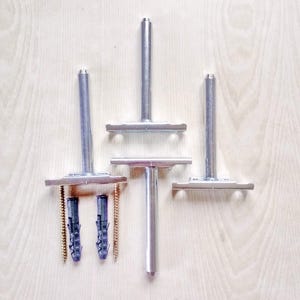 May include: An overhead shot shows three metal shelf supports with a T-shaped design, along with two plastic wall anchors and two screws. The supports are silver and appear to be made of metal. The background is a light wood grain.