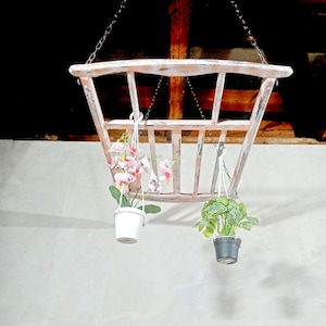 Puede incluir: Un macetero colgante de madera blanca con dos plantas. Una planta tiene flores rosas y la otra tiene hojas verdes. El macetero está colgado del techo con cadenas.