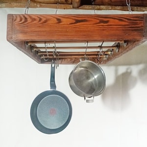 May include: A wooden pot rack suspended from chains, featuring copper rods and hooks. A black frying pan and a silver pot hang from the rack. The wood has a warm, reddish-brown tone.