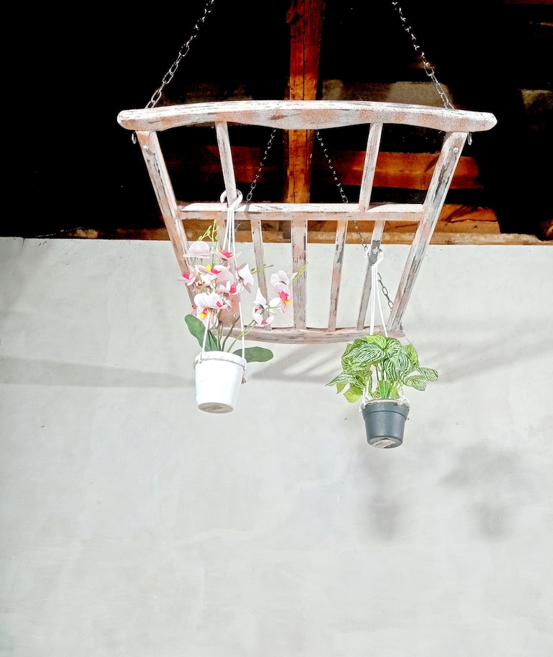 May include: A white wooden hanging planter with two plants. One plant has pink flowers and the other has green leaves. The planter is hanging from the ceiling with chains.