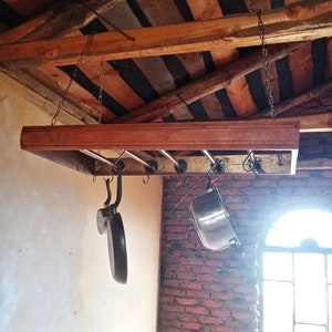 Puede incluir: Un estante de madera para ollas con tuberías metálicas y ganchos que cuelgan del techo. El estante sostiene una variedad de ollas y sartenes, incluyendo una olla grande de plata y una sartén negra.