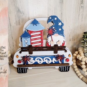 Könnte beinhalten: Ein Holz-Zwerg mit einem rot-weiß-blauen Hut hält ein Feuerwerk und fährt in einem weißen Pickup-Truck. Der Truck ist mit rot-weiß-blauen Feuerwerkskörpern verziert und das Wort "BOOM" steht auf der Rückseite.