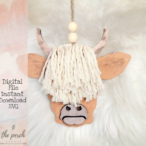 May include: A wooden cutout of a Highland cow head with a white fringe of yarn attached to the top. The cow has a brown and white painted design and a light brown nose. The cutout is hanging from a string with two wooden beads.