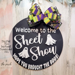 May include: A black round wooden sign with white text that reads "Welcome to the Sheet Show! Hope you brought the boos!"  Two white ghost silhouettes are on the sign. A green and purple bow is attached to the top of the sign.