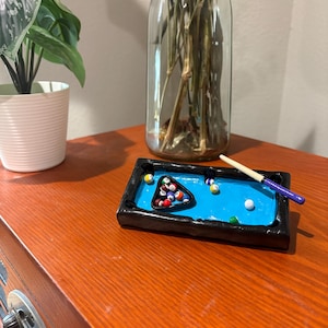 Op de afbeelding: Een miniatuur pooltafel met een blauw speelveld, zwart frame en kleurrijke poolballen. Een kleine keu met een paarse handgreep ligt op de tafel. De tafel is een decoratief item.