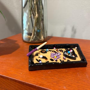 Op de afbeelding: Een rechthoekige asbak met een zwarte rand en een luipaardprint interieur. De asbak heeft een miniatuur pooltafelontwerp met biljartballen, een driehoekig rek en een keu. De tekst "The Cheetah Girls" is zichtbaar.