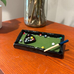 Op de afbeelding: Een miniatuur asbak in de vorm van een pooltafel. De asbak heeft een groen vilt oppervlak, zwarte randen en een set miniatuur poolballen gerangschikt in een driehoek. Een kleine keu ligt op de tafel.