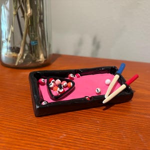 Op de afbeelding: Een miniatuur pooltafel met een roze vilt oppervlak en zwart frame. De set bevat een rek met biljartballen, diverse losse ballen en twee miniatuur keuen met blauwe en rode uiteinden. Een decoratief item.