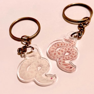 May include: Two clear acrylic keychains featuring a coiled snake design. One snake is light gray with subtle spots, and the other is light pink with darker pink spots. Both keychains are attached to circular bronze-colored metal key rings.