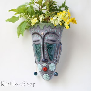 Könnte beinhalten: Ein Keramik-Wandtopf in Form eines Gesichts mit geschlossenen Augen, einem roten Mund und blauen und grünen geometrischen Mustern. Der Topf ist mit gelben und weißen Blumen gefüllt.