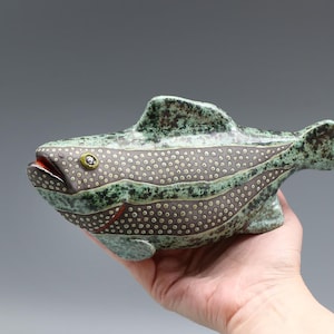 May include: A ceramic fish sculpture with a speckled gray body, green accents, and a red mouth. The fish has a textured surface and is held in a hand. The sculpture is approximately 10 inches long.