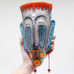 May include: A ceramic wall hanging in the shape of a face with a blue, orange and grey colour scheme. The face has closed eyes, a small nose and a large mouth. The hanging has red tassels.