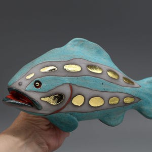 Könnte beinhalten: Eine Keramikfischskulptur mit einem gesprenkelten türkisfarbenen Körper, einem grauen Seitenstreifen, ovalen Blattgoldakzenten und einem leuchtend roten Maul. Der Fisch hat ein sichtbares Auge mit goldener Iris und dunkler Pupille.