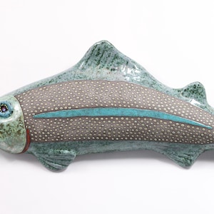 Peut inclure: Une sculpture de poisson en céramique dans les tons de vert, gris et bleu. Le poisson a un corps texturé avec un motif pointillé et une bande turquoise. La bouche est rouge et les yeux bleus.