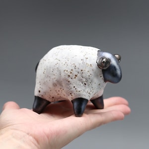 Puede incluir: Una pequeña figura de oveja de cerámica con un cuerpo blanco texturizado con motas marrones, una cabeza lisa de color metal oscuro y cuatro patas cortas oscuras. La oveja tiene dos pequeños ojos saltones y una forma compacta de cuatro patas.