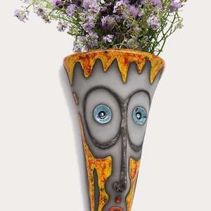 Può includere: Un vaso da parete in ceramica a forma di viso dipinto di giallo, arancione e rosso. Il vaso ha grandi occhi blu e una piccola bocca rossa. Il vaso è pieno di fiori viola.