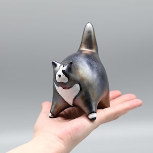 Peut inclure: Figurine de chat en céramique avec une finition gris métallisé et bronze. Le chat a des marques blanches sur la poitrine et le visage, avec des accents blancs sur les pattes. Le chat est dans une pose ludique avec la queue levée.