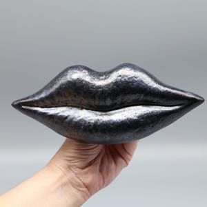 Op de afbeelding: Een metallic zwarte lippensculptuur, vastgehouden in een hand. De lippen hebben een glanzend, getextureerd oppervlak met een zilveren glans. Het kunstwerk is een decoratief stuk.