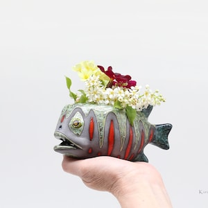 Könnte beinhalten: Ein Keramik-Fisch-Pflanztopf mit grüner, grauer und roter Glasur. Der Topf ist mit weißen und rosa Blumen gefüllt.