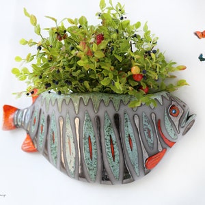 Könnte beinhalten: Ein Keramik-Fisch-Pflanztopf mit einem grün-grau-orangenen Design. Der Topf ist mit einer grünen Pflanze mit kleinen roten Beeren gefüllt. Zwei Schmetterlinge fliegen in der Nähe des Topfes.
