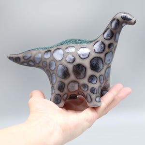 Könnte beinhalten: Eine Keramik-Dinosaurierfigur mit einem gesprenkelten Muster in Grau und Weiß und einem türkisfarbenen Akzent auf der Rückseite. Der Dinosaurier steht auf einer Hand.