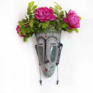 Könnte beinhalten: Eine Keramik-Wandvase in Form eines Gesichts mit grünen und grauen Akzenten. Die Vase hat kleine Löcher zum Aufhängen und ist mit kleinen weißen Punkten verziert. Die Vase ist mit rosa Blumen gefüllt.