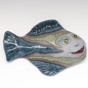 Peut inclure: Une décoration murale en céramique en forme de poisson avec un motif rayé bleu, vert et blanc. Le poisson a un visage blanc avec des yeux verts et une bouche rouge.