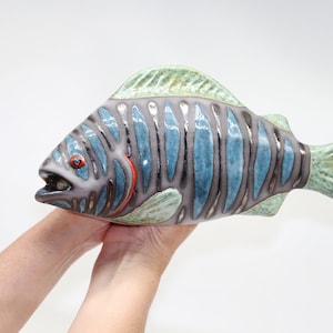 Könnte beinhalten: Eine Keramik-Fischskulptur mit blauen, grünen und grauen Streifen. Der Fisch hat ein rotes Auge und einen orangefarbenen Mund.