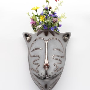 May include: A gray ceramic wall planter shaped like a cat's face with a brown outline. The planter has a small opening at the top and is filled with a bouquet of wildflowers, including yellow, purple, and pink flowers. A red butterfly is flying above the flowers.