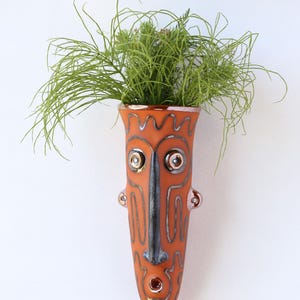 May include: An orange ceramic wall vase with a tribal mask design. The vase has a long, narrow shape with a face-like design, including eyes, a nose, and a mouth. The vase is filled with green plants. The vase is approximately 10 inches tall.