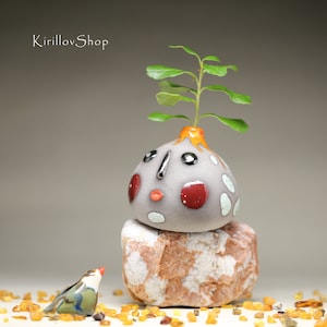 May include: A small gray ceramic planter with a face and colorful accents, topped with a green plant. The planter is sitting on a brown rock with a small bird figurine nearby.