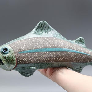 May include: A ceramic fish sculpture with a speckled brown body, teal stripe, and teal fins. The fish has a red mouth and blue eyes. The sculpture is held in a hand against a gray background.