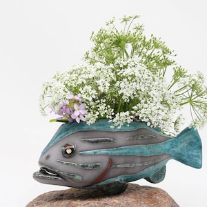 Può includere: Un vaso da fiori in ceramica a forma di pesce con una glassa verde acqua e grigia, che presenta un design dettagliato di squame e pinne. Il vaso è pieno di fiori bianchi e steli verdi.