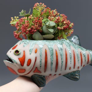 May include: A ceramic fish-shaped planter with a white, orange, and teal design, holding a vibrant arrangement of red and green flowers. The planter has a detailed, textured finish and is held by a hand.