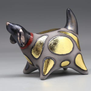 Könnte beinhalten: Eine Keramik-Hundefigur mit einem matten grauen Körper, verziert mit metallischen Blattgoldflecken sowie einem dunklen, irisierenden Kopf und Schwanz. Der Hund trägt ein dünnes rotes Halsband und steht auf vier kurzen, stabilen Beinen.