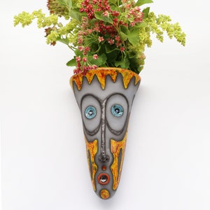 Könnte beinhalten: Eine Keramik-Wandvase in Form eines Gesichts mit gelber, orangefarbener und roter Farbe. Die Vase hat blaue Augen und einen roten Mund. Sie hält einen Strauß rosa und grüner Blumen.