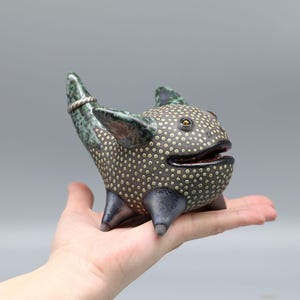 Könnte beinhalten: Eine Keramikfigur eines skurrilen Wesens mit einem gefleckten schwarzen und grünen Körper, einem weit geöffneten Maul und einem Schwanz mit einem silbernen Ring. Das Wesen hat vier Beine und ein Paar Flügel.