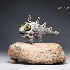 May include: A small, white ceramic sculpture of a stylized creature with a spiky back, yellow eyes, and a gray and white patterned body. The creature is standing on a brown rock.