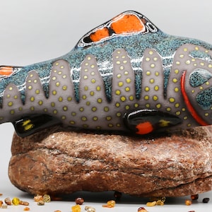 Könnte beinhalten: Handbemalte Keramik-Fischfigur mit einem blau, grau und weiß gesprenkelten Körper, orangefarbenen und gelben Flossen und schwarzen Akzenten. Der Fisch sitzt auf einem braunen Stein.
