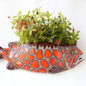 May include: A ceramic fish-shaped planter with a grey body and orange spotted pattern. The fish has a brown tail and fins, and a wide opening filled with green foliage, red berries, and dark blue berries. The planter is on a white background.