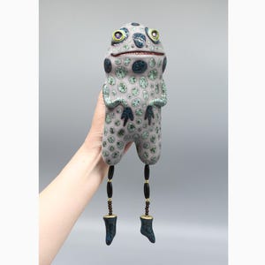 May include: A ceramic frog figurine with green and black spots, yellow eyes, and brown and black beaded legs. The frog is standing on a grey background.